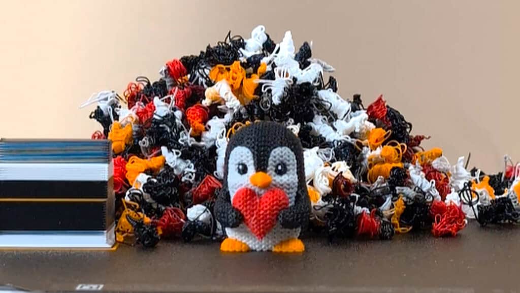 Close up of a multi color print of a penguin holding a red heart on a bambu lab a1 with a pile of purged filament in the background