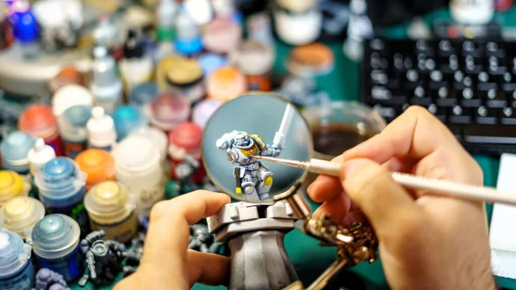 Looking through a magnifying glass as a man paints a warhammer miniature that was resin 3d printed. There are paints, a keyboard, and other miniatures in the background